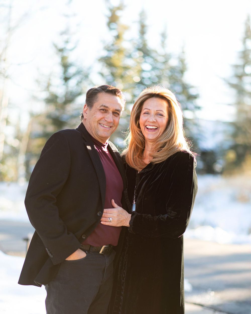 A couple in formal winter attire poses together in a snowy mountain setting.