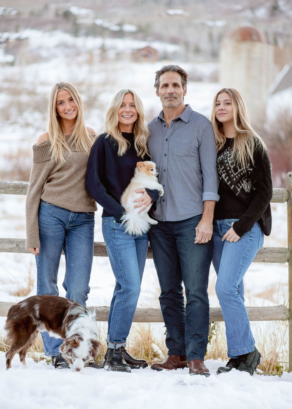 Family outdoor portrait with dogs in snowy setting against rustic fence and mountain backdrop.