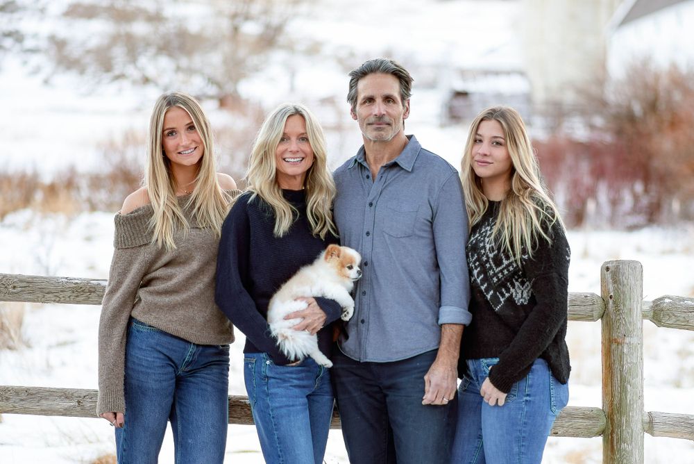 A family group with a small white dog poses along a fence in a winter landscape.