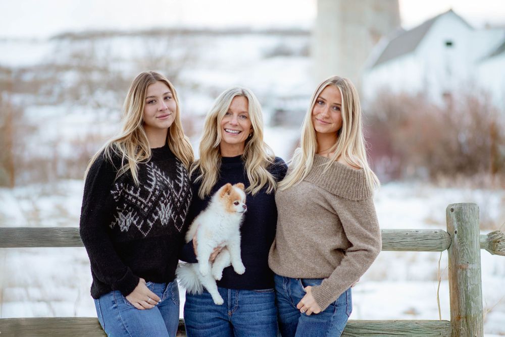 Three people in sweaters hold a small white dog while posing by a fence in winter.