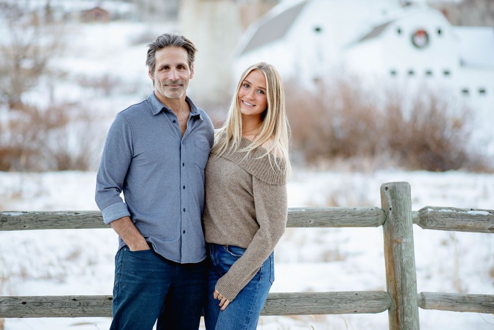 Couple posing together against snowy winter landscape with wooden fence.