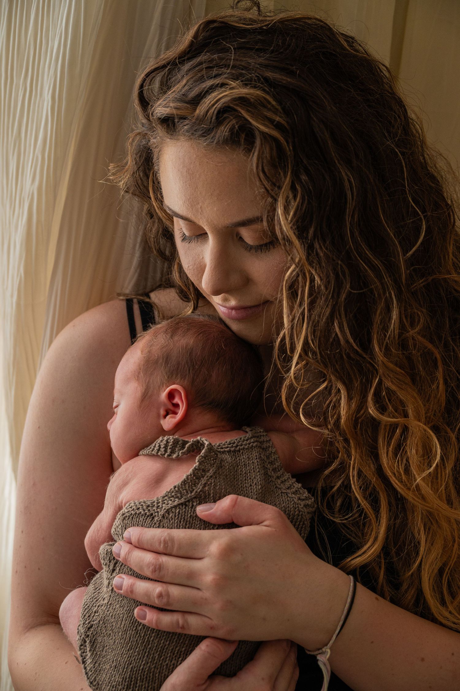 Mother with long curly hair holding sleeping newborn baby in knit outfit.