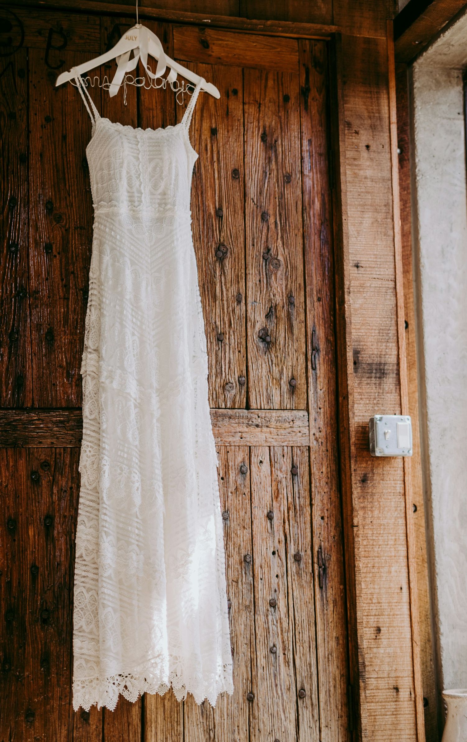 A delicate white lace wedding dress hangs from a decorative hanger against a rustic wooden door background.