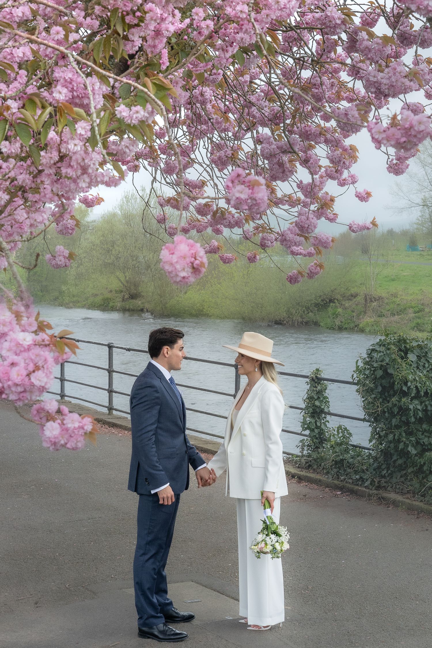 A couple stands beneath blooming cherry blossoms along a riverside path.