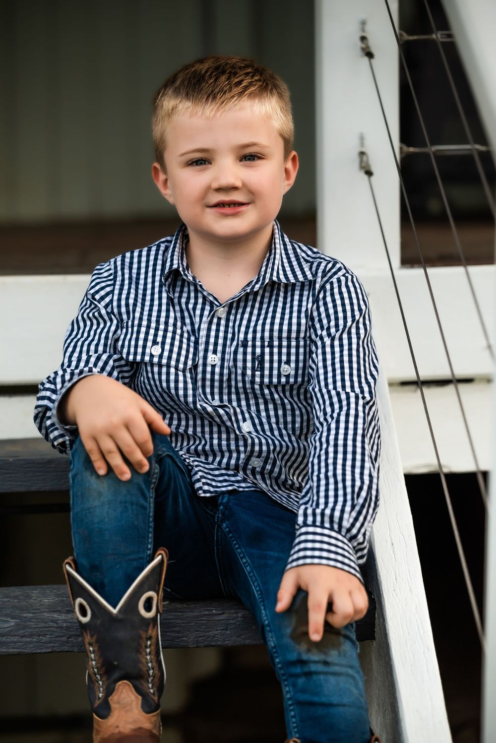 Young boy in plaid shirt and cowboy boots sitting on white porch steps.