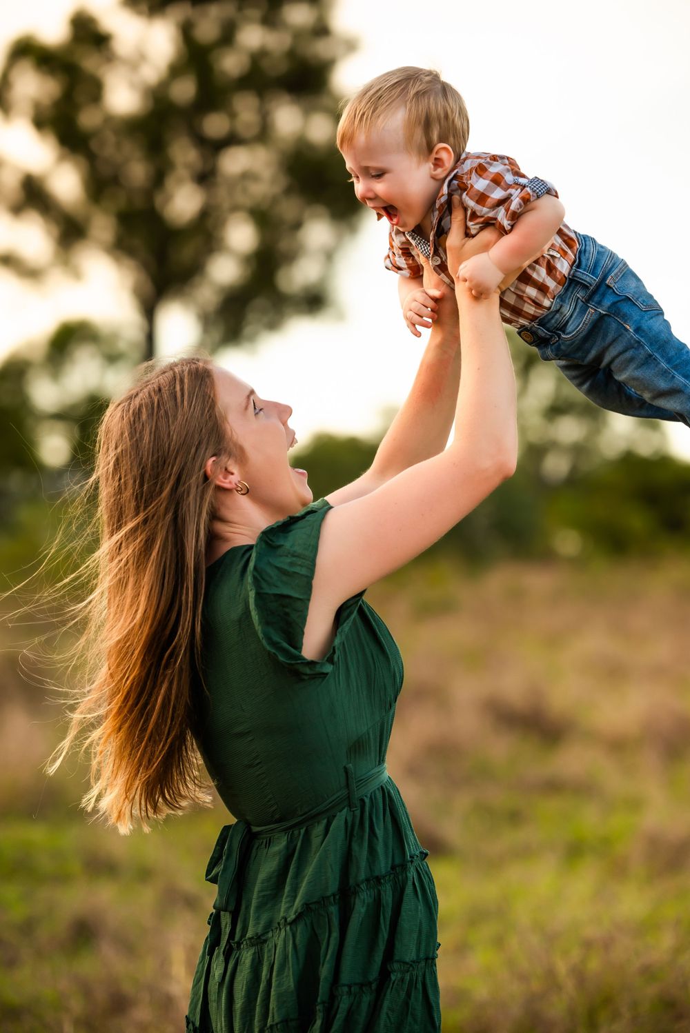 Woman in green dress lifts and plays with laughing baby outdoors at sunset.