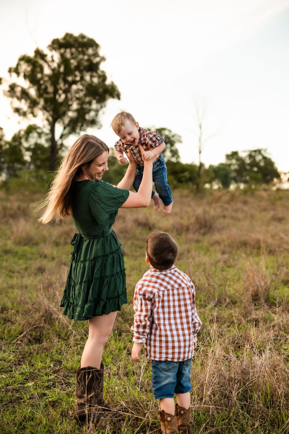 Mother lifts baby high while another child watches in a sunlit field.