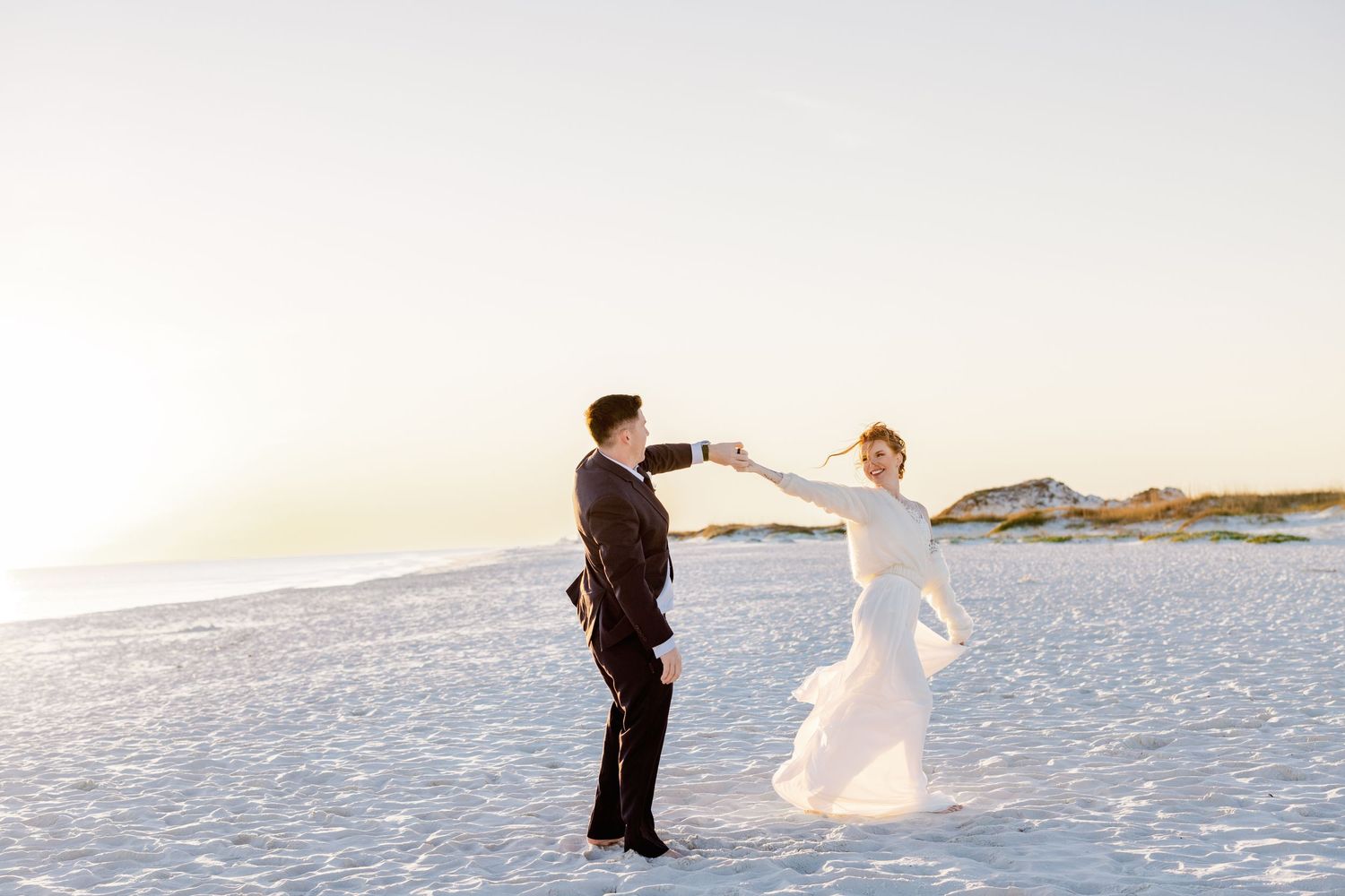 A couple in wedding attire dance together on a pristine white sandy beach at sunset.