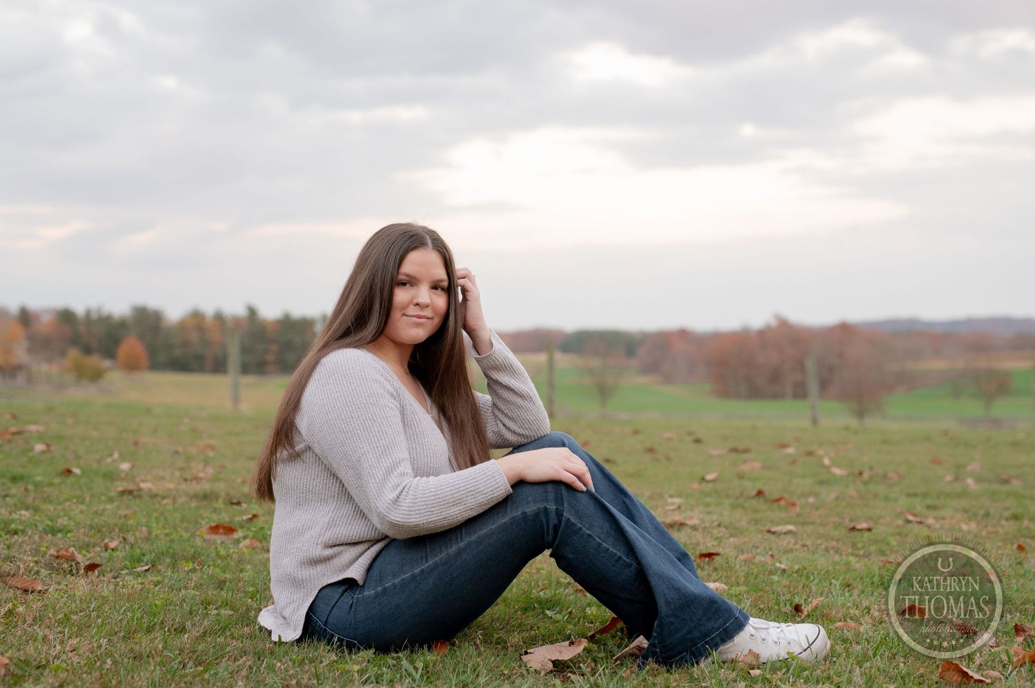 Person sits in field wearing gray sweater with cloudy autumn sky above.