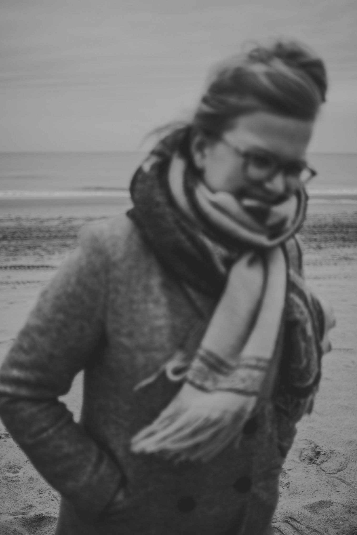 Zwart wit foto van een vrouw met haar in een knot, bril op en sjaal om op het strand.
