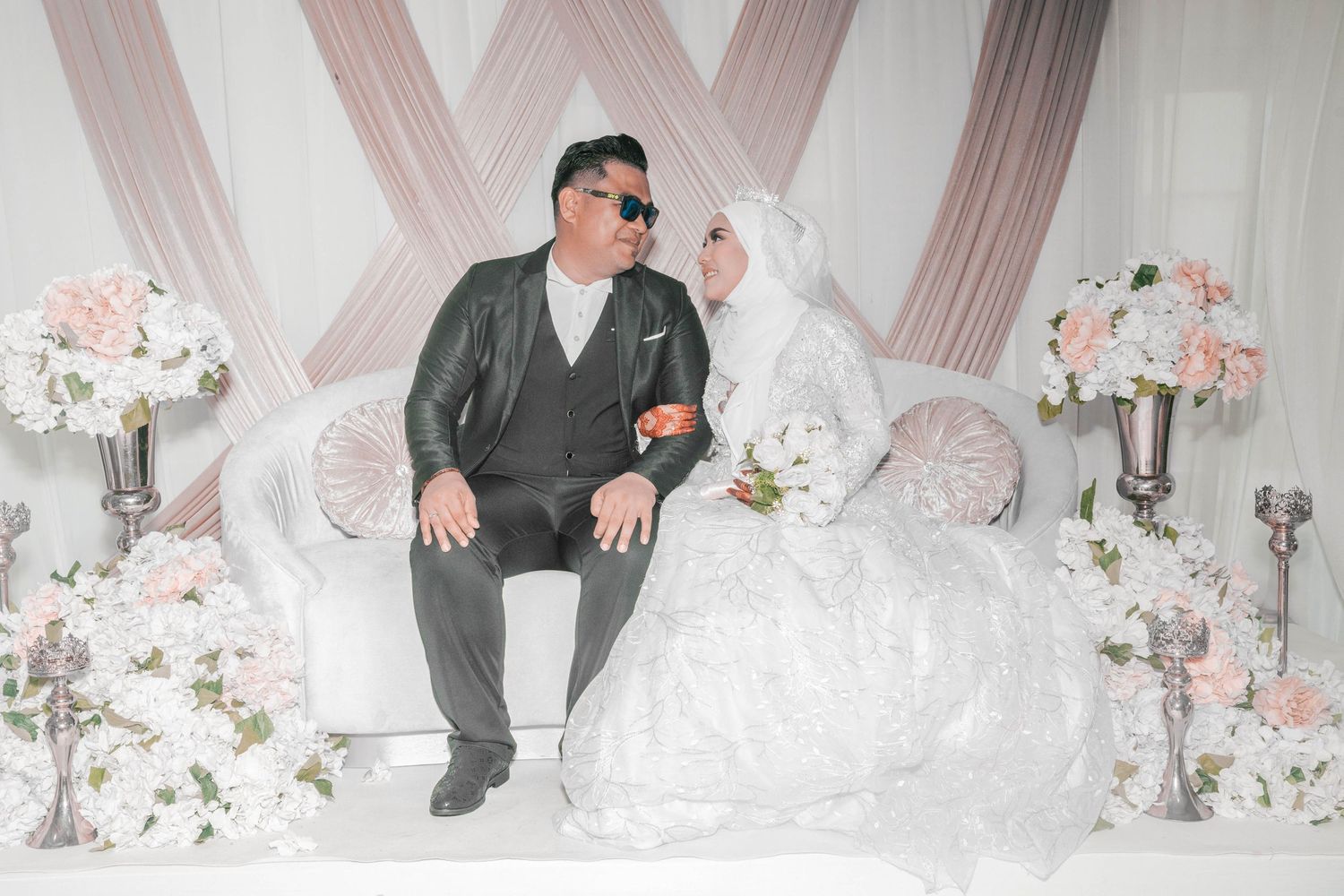 Wedding couple seated on decorated white stage with floral arrangements and soft pink drapery.