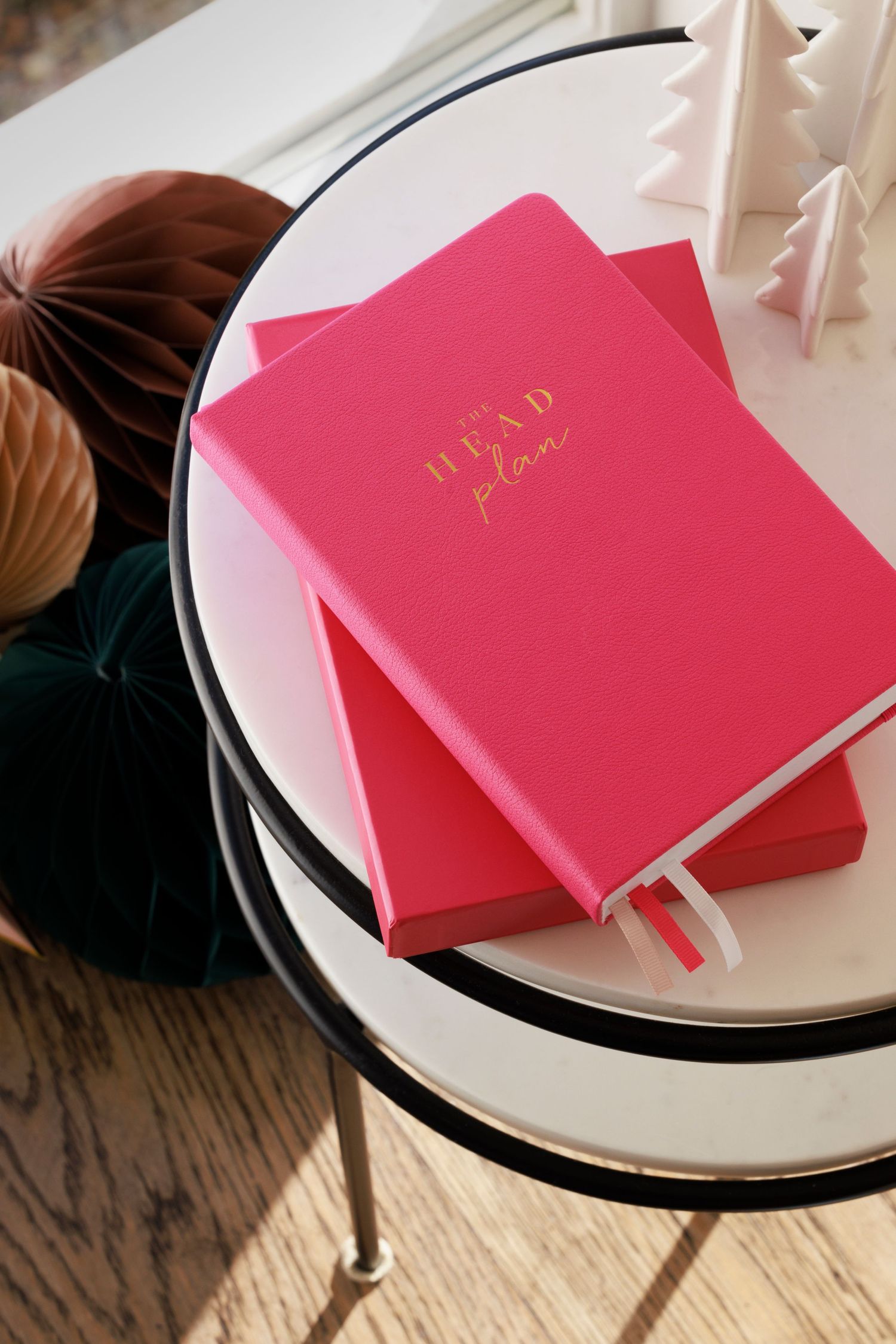 Pink hardcover notebooks with gold text resting on white plates against a wooden surface with decorative elements.