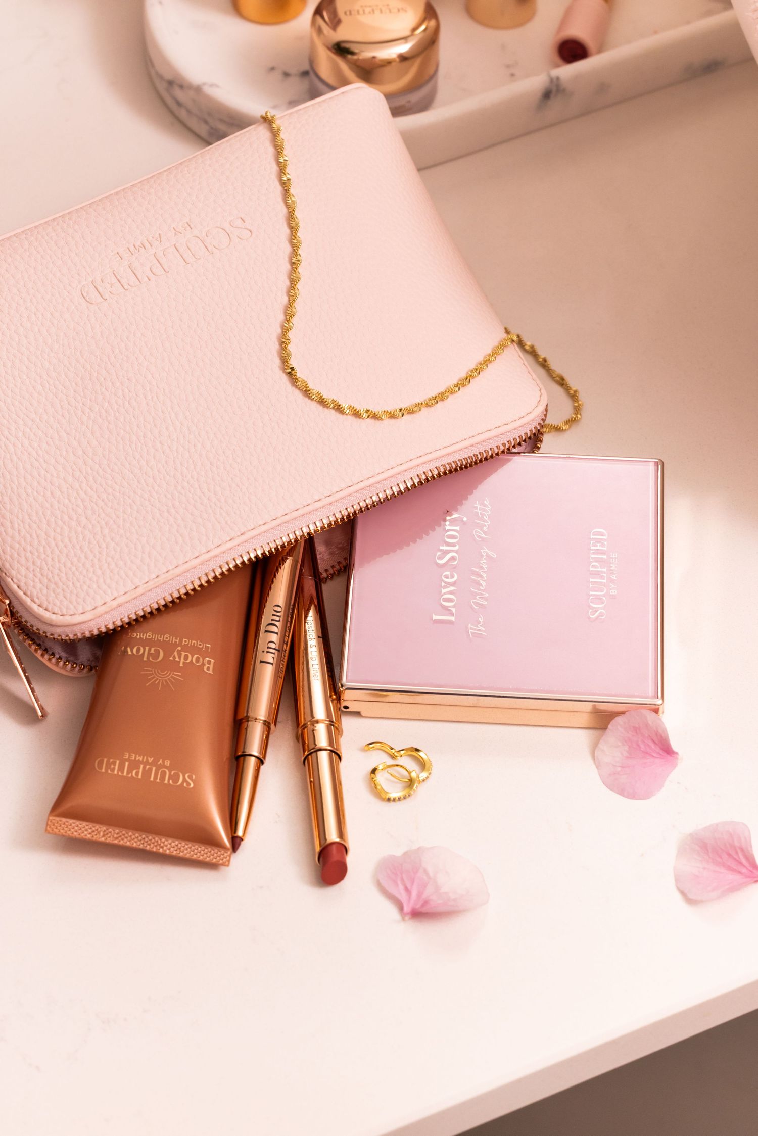 Pink leather wallet with gold chain and makeup products arranged on white surface with rose petals.