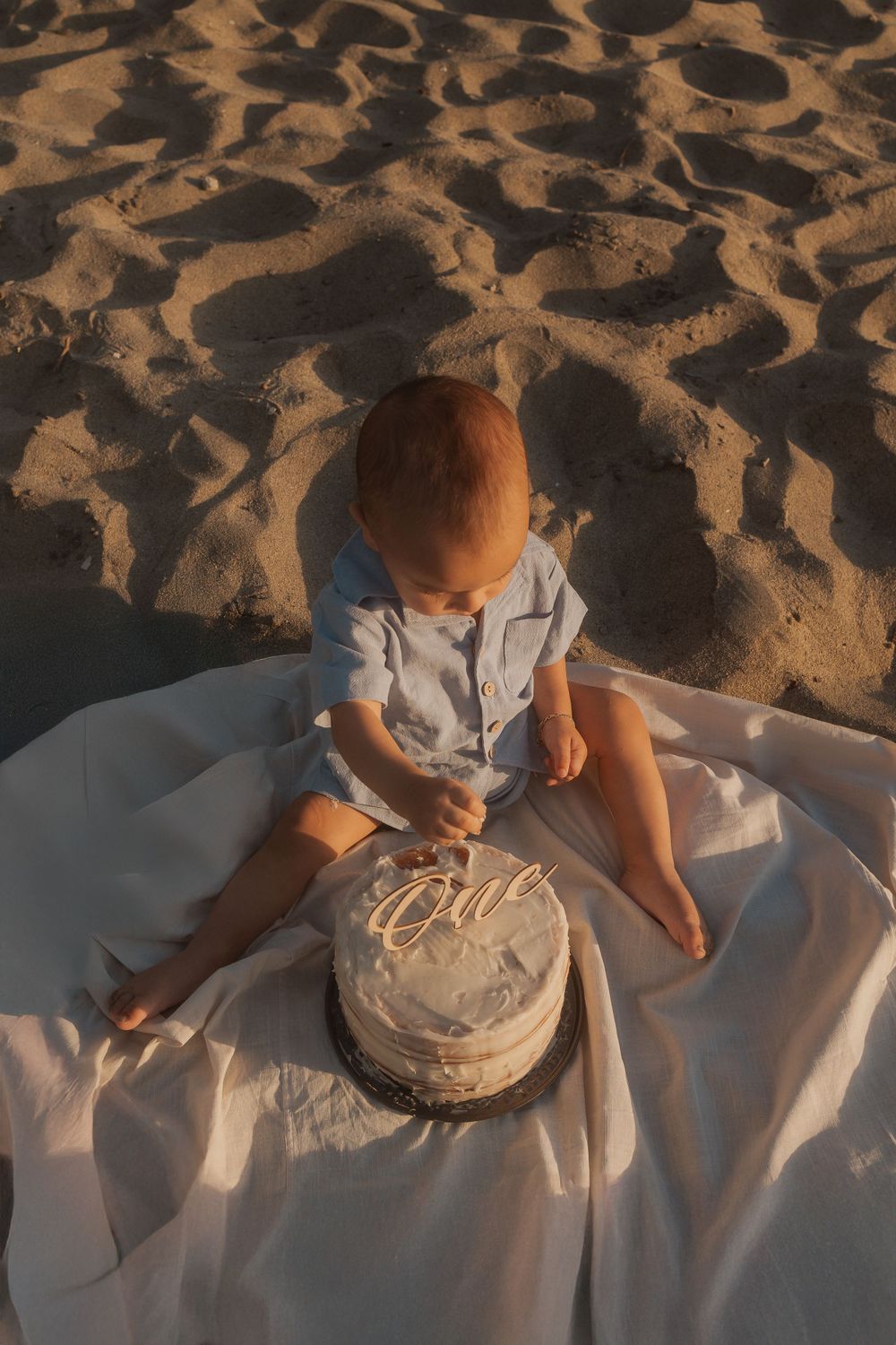 Top view of toddler sitting on white blanket with cake on textured beach sand.