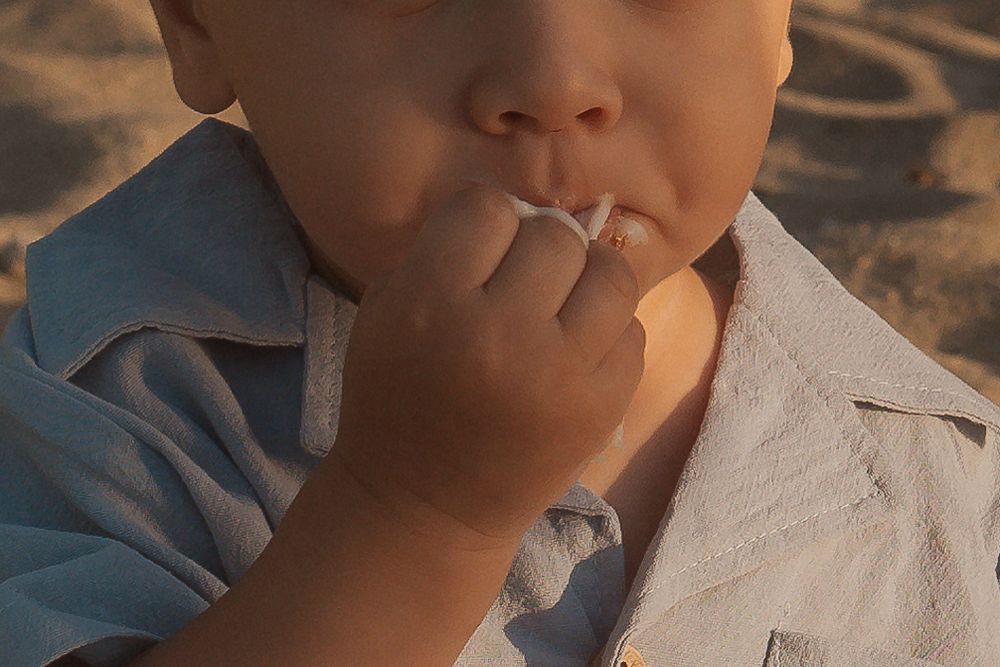 Small toddler reaching towards mouth with frosting while sitting on beach blanket.