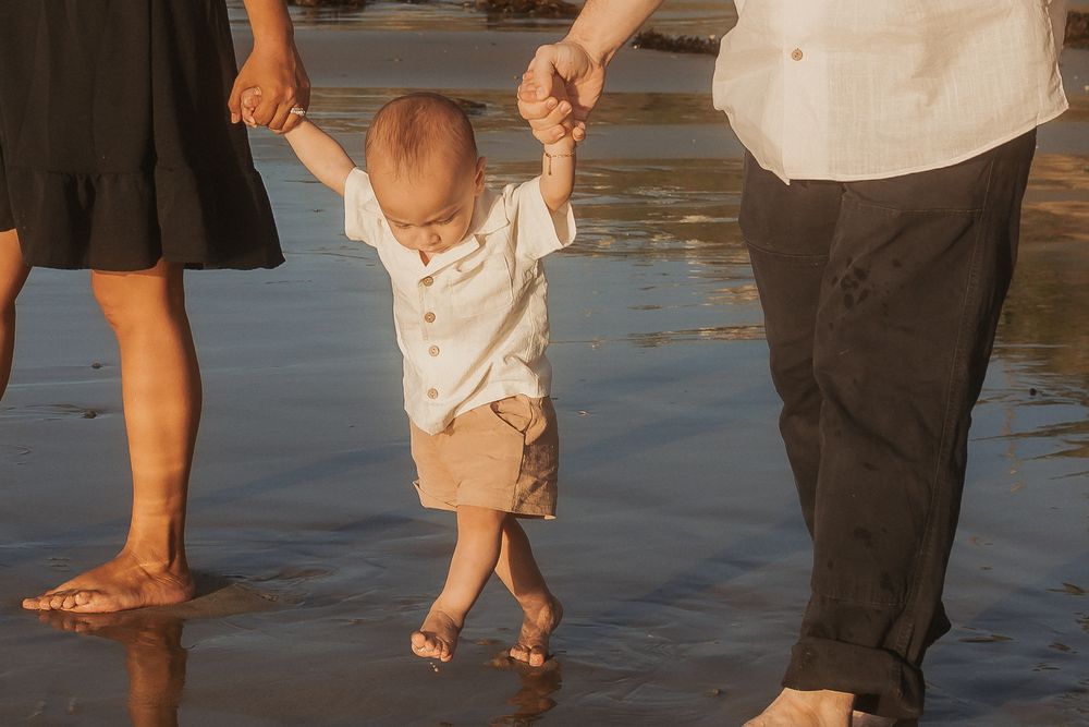 Young child walking on wet sand at the beach held by two people.