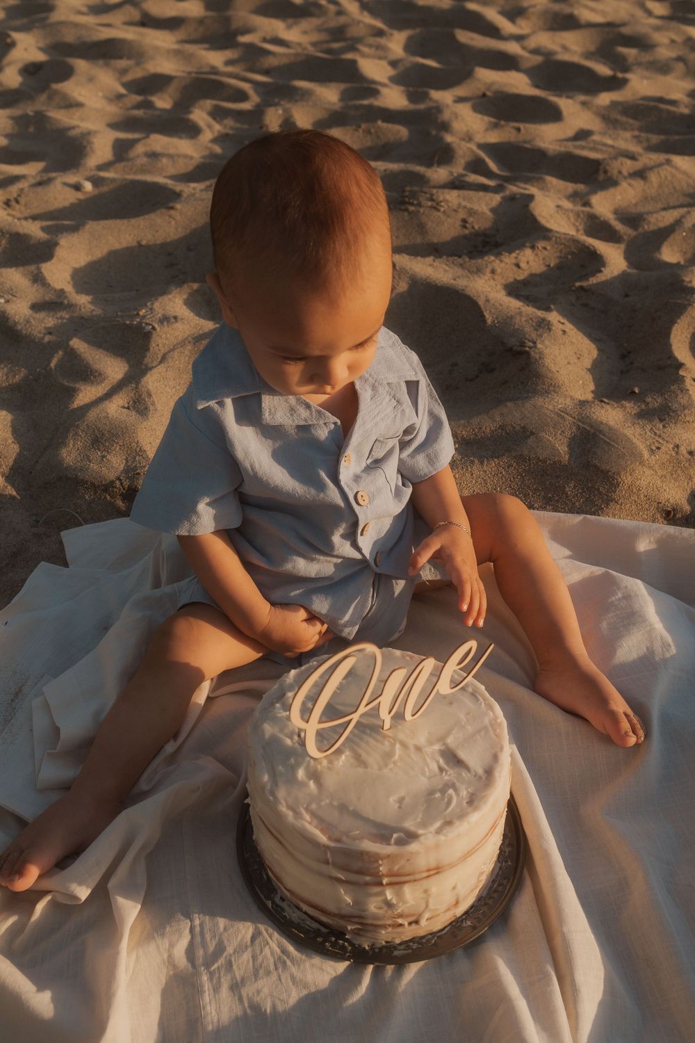 A small child in blue sits on a white blanket on sand with a decorated cake showing 'One'.