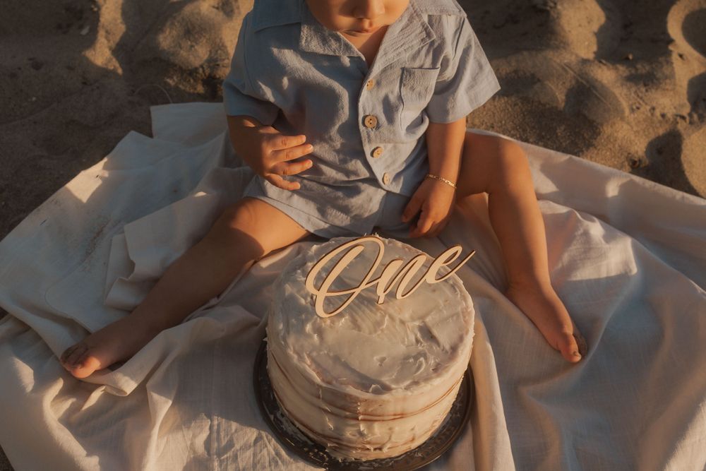 A toddler with decorated white birthday cake sits on a blanket at the beach during golden hour.