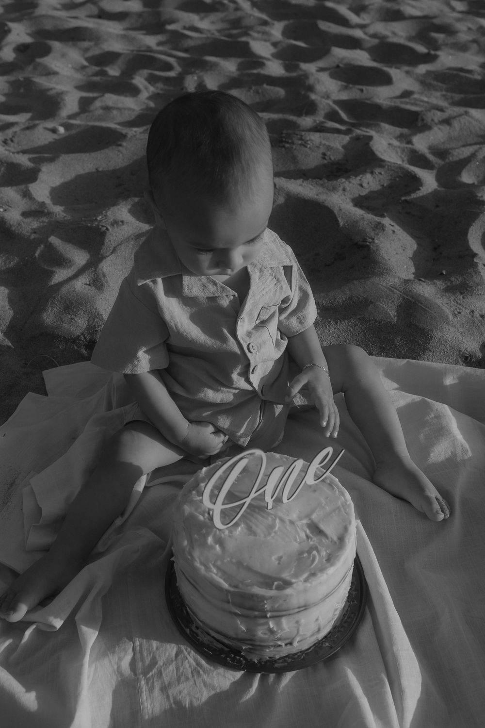 Black and white photo of toddler with smash cake on beach blanket.