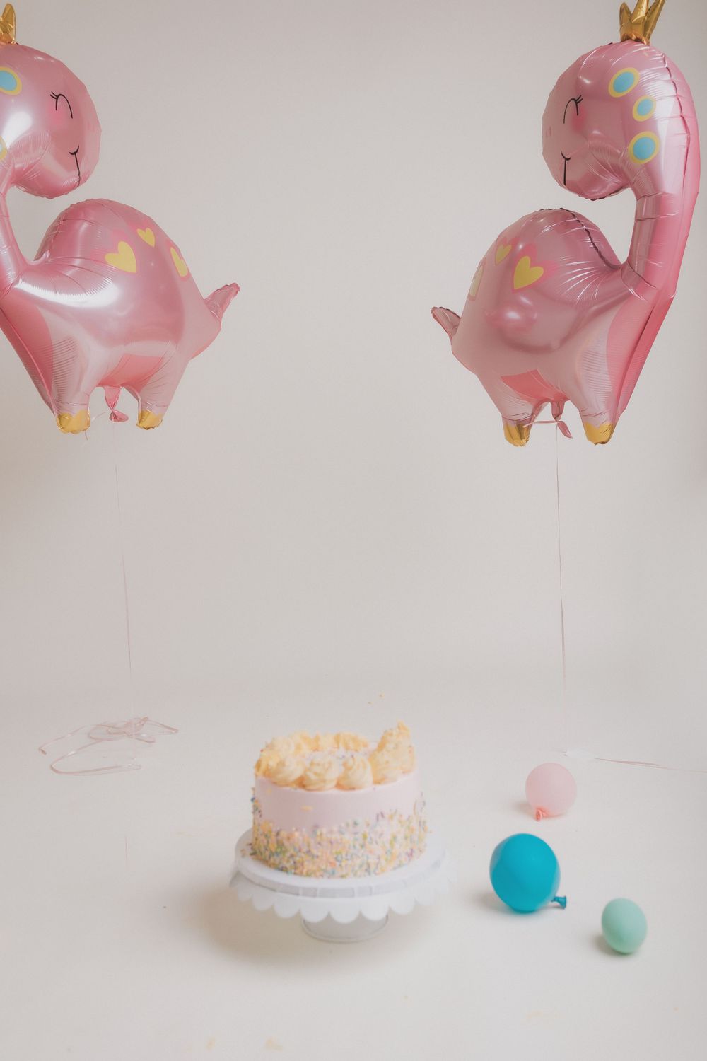 Two pink dinosaur balloons hover above a white birthday cake with pastel decorations.