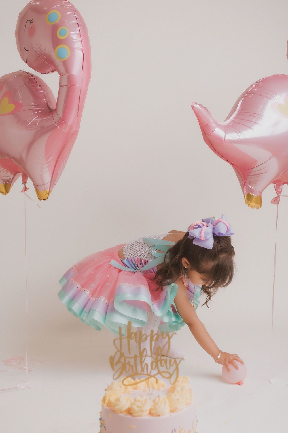 A festive birthday setup with pink dinosaur balloons and a decorated cake on white pedestal.