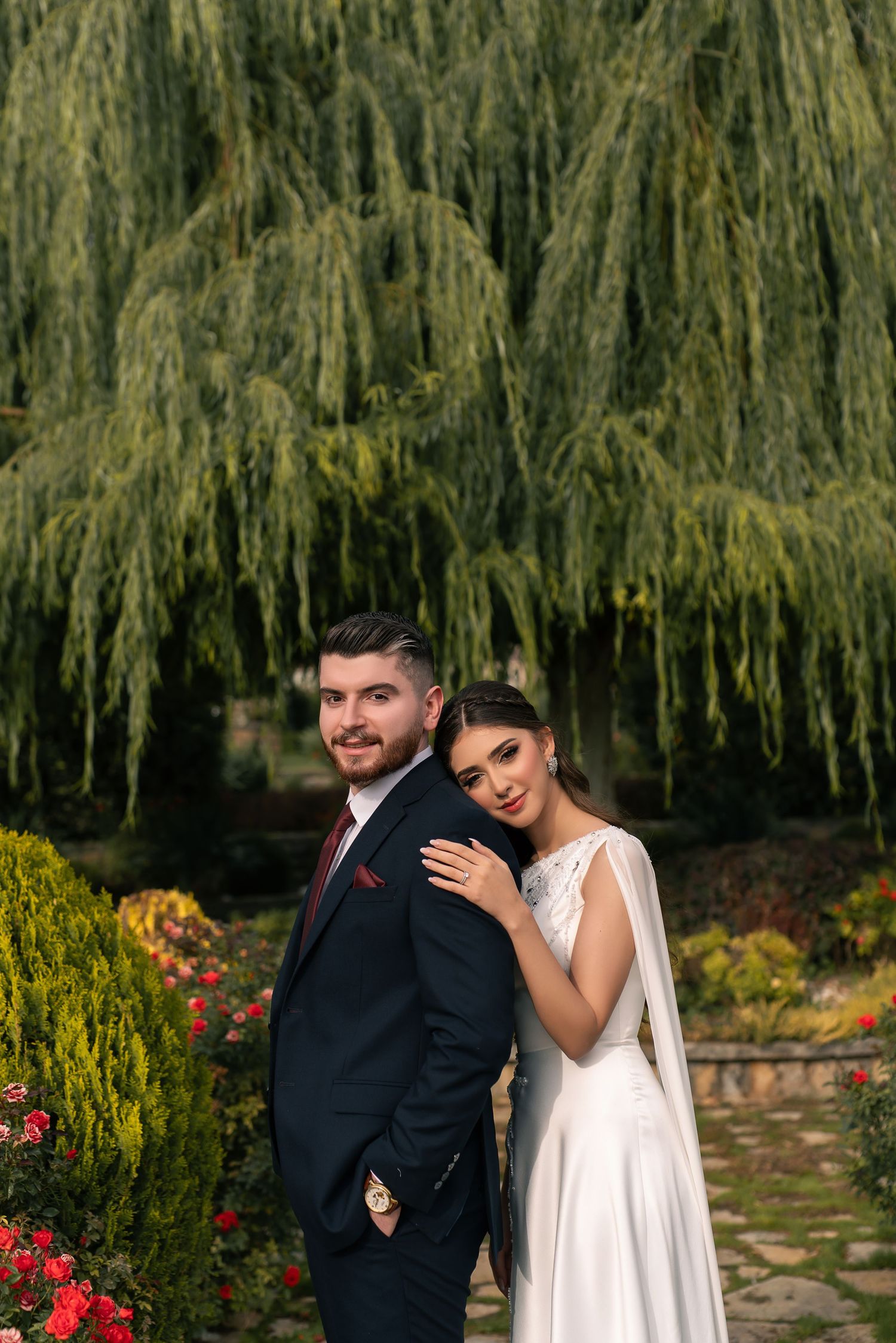 A romantic wedding couple poses in a garden with cascading willow trees and colorful flowers in the background.