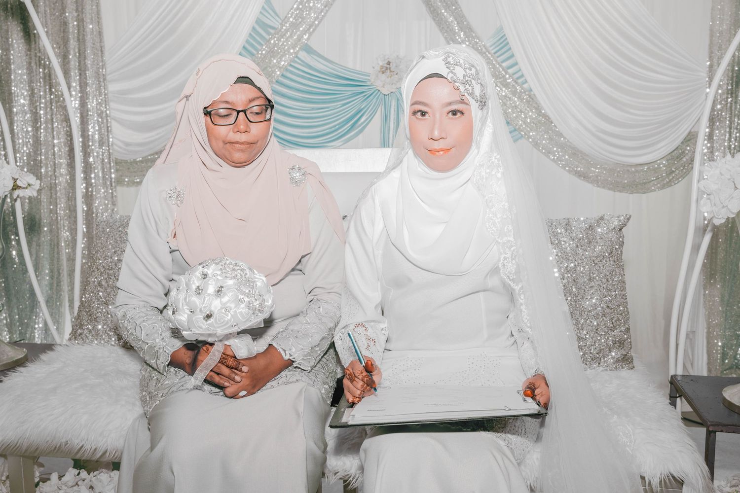 Two people in white attire seated during a traditional wedding ceremony.