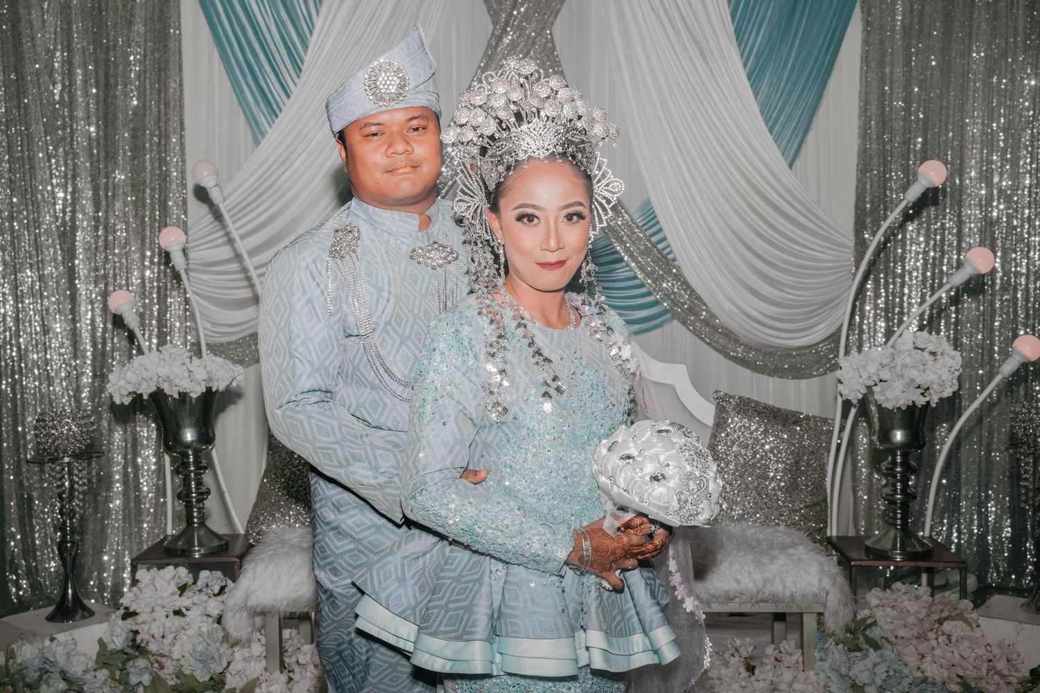 Formal portrait of a couple in matching silver outfits against a decorated backdrop with teal and white draping.