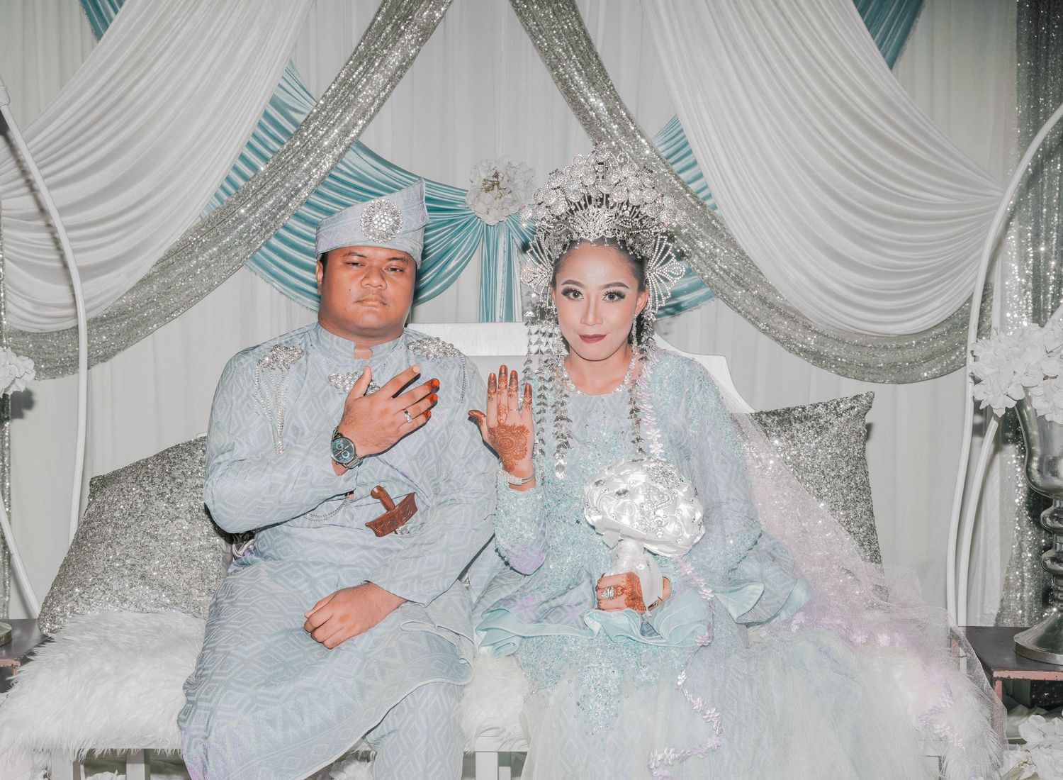 Couple in matching silver attire seated together at formal event with teal and white drapery.