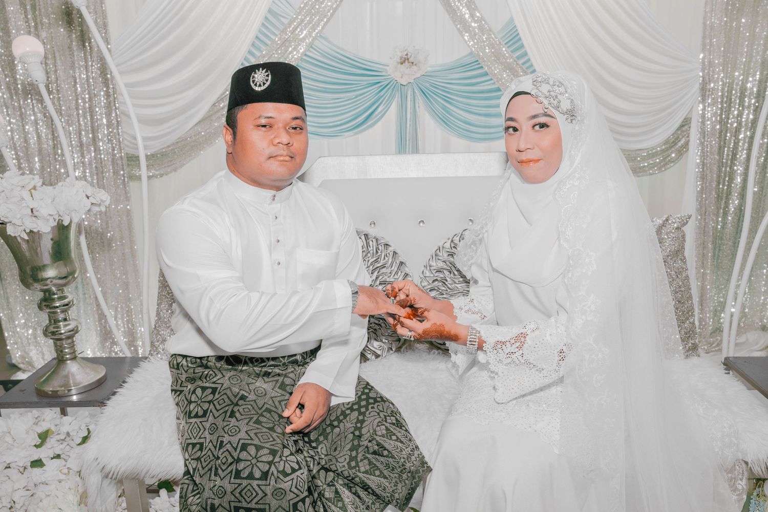 Wedding couple in traditional Malay attire seated on white and silver decorated stage.