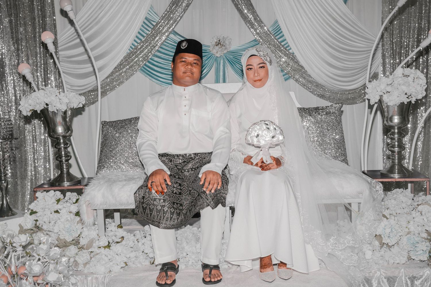 Newlyweds in traditional Malay wedding attire seated in silver and white decorated setting.