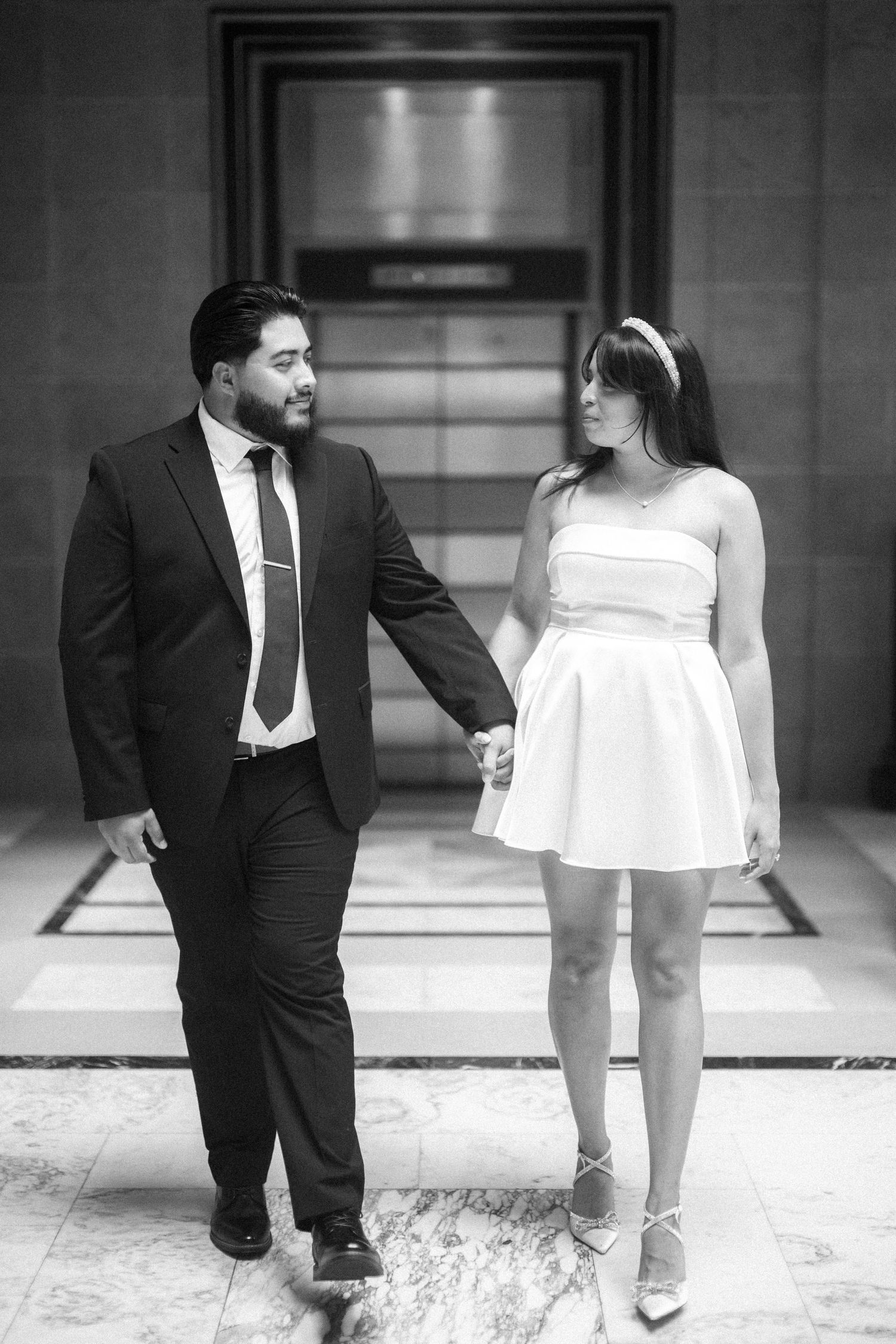 A couple in formal attire holds hands while walking together, with her in a white dress and him in a dark suit.