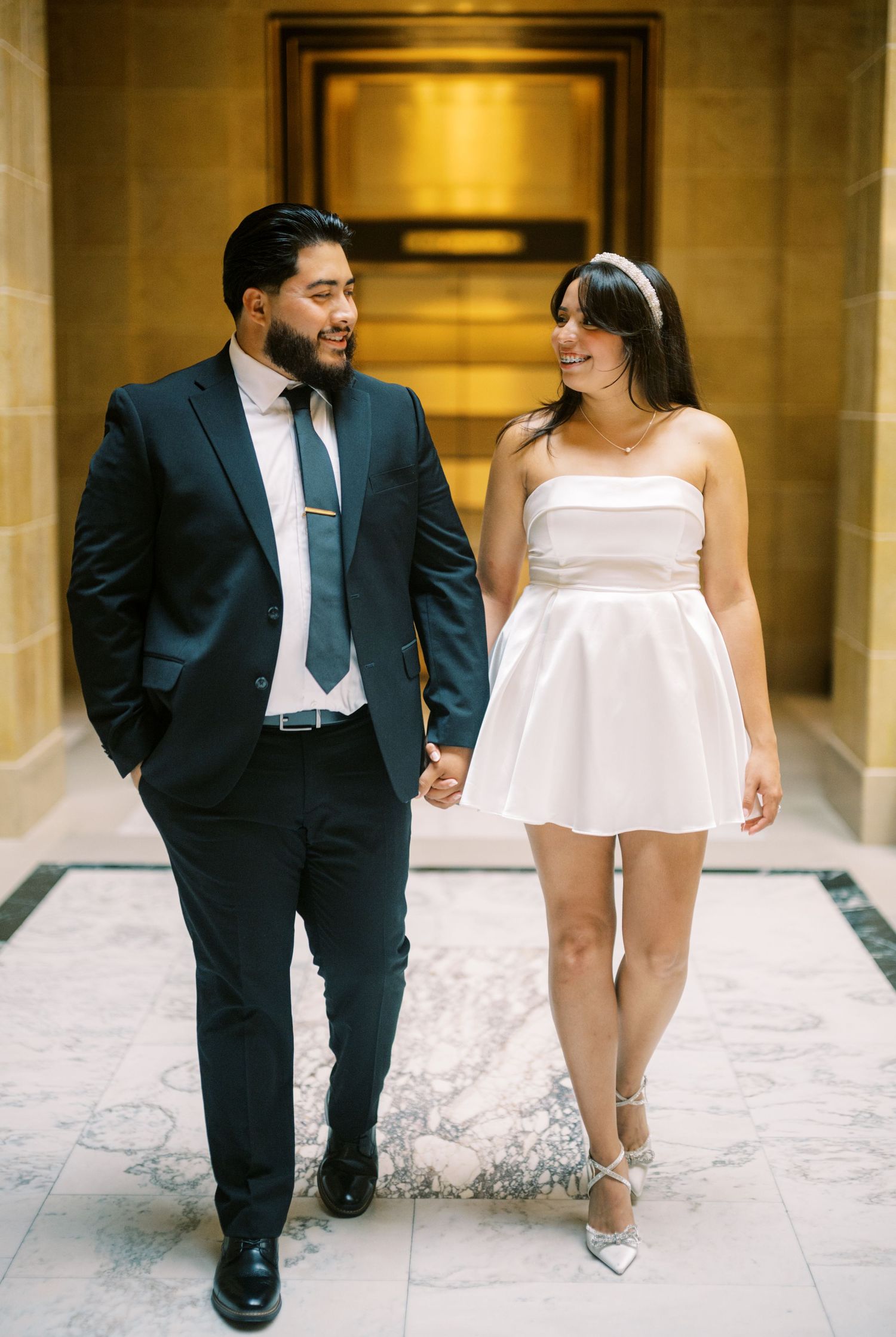 Couple walking together in formal attire - a navy suit and white dress - through an elegant stone hallway.