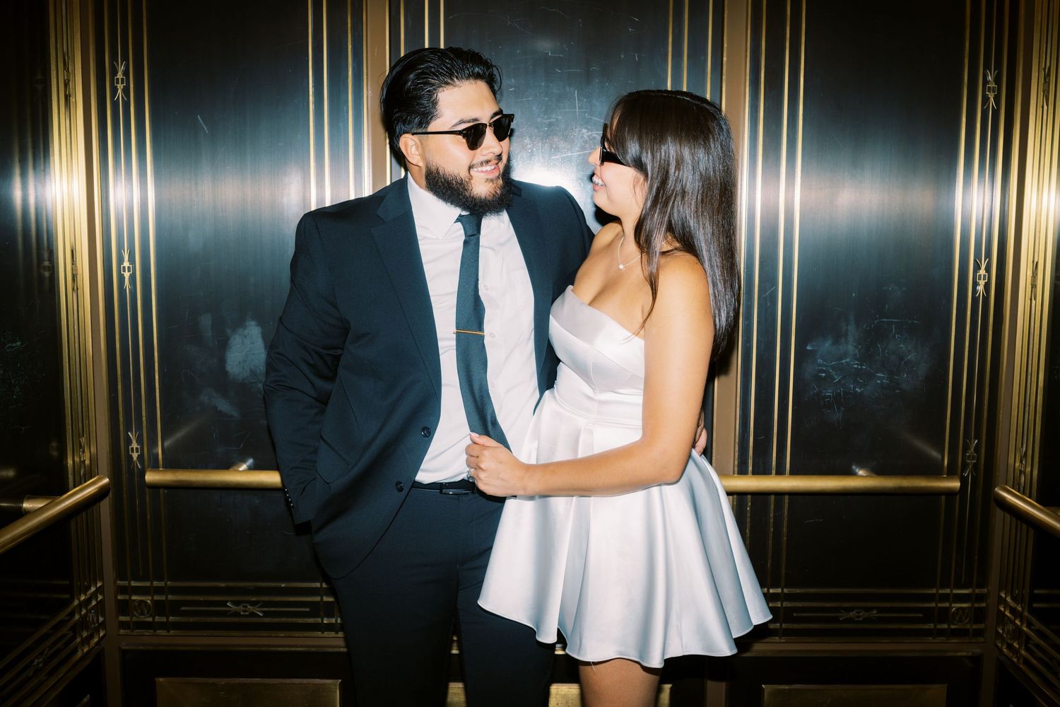 A couple in formal attire poses together intimately in an elegant gold-trimmed elevator.