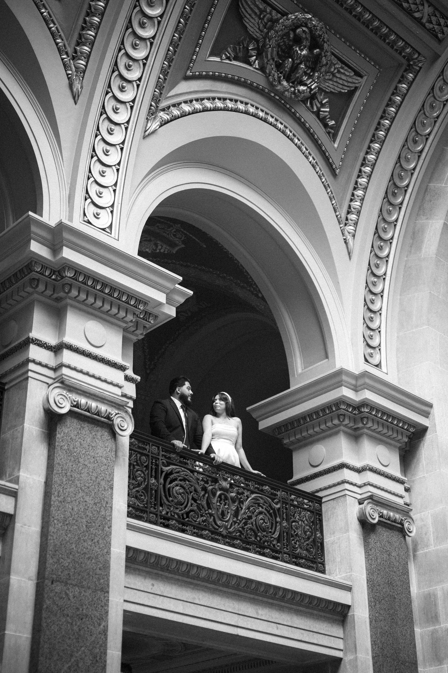 A couple stands together on an ornate balcony with classical architecture featuring detailed arches and moldings.