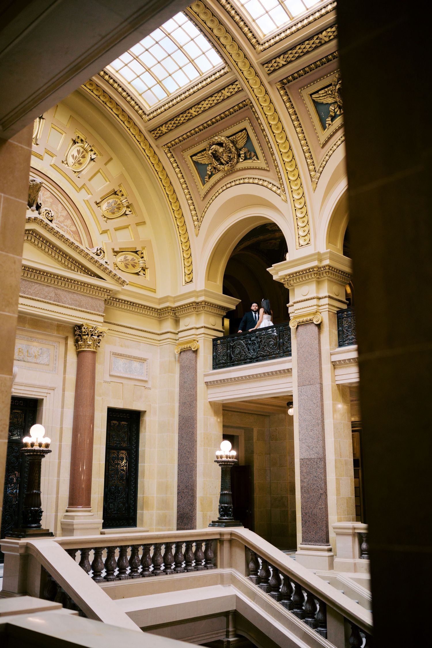 Ornate neoclassical interior staircase with cream marble columns, arched ceilings, and decorative skylight in historic building.