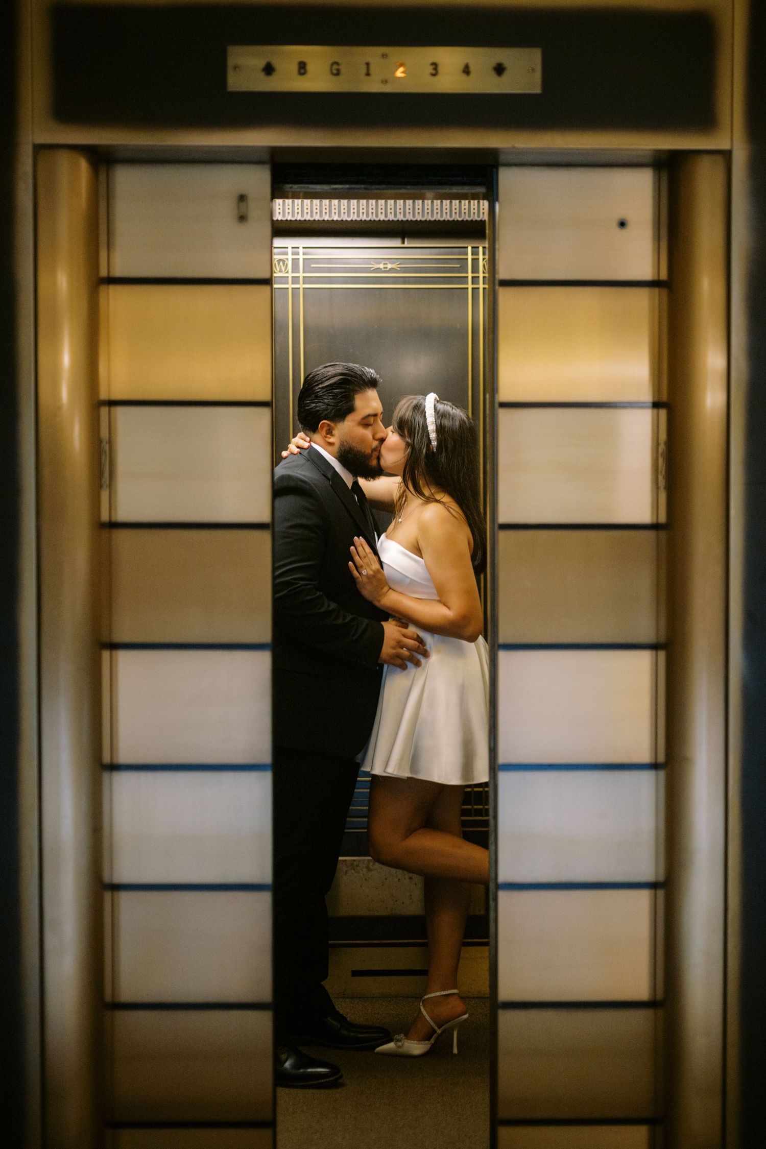 A couple shares a romantic moment inside an elevator, captured through the partially open doors at their floor.