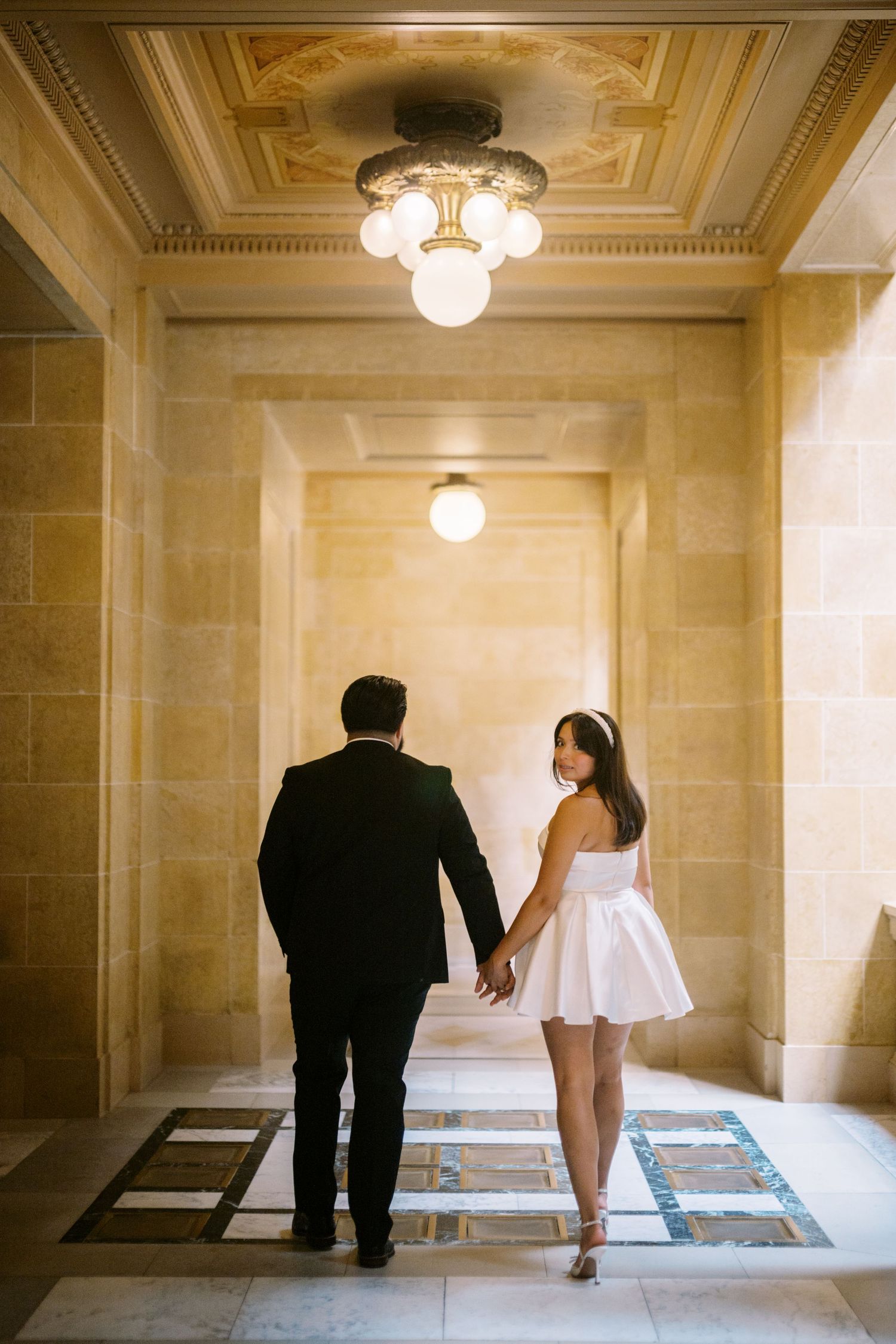 A couple holding hands walking down an elegant hallway with marble walls and ornate lighting fixtures.