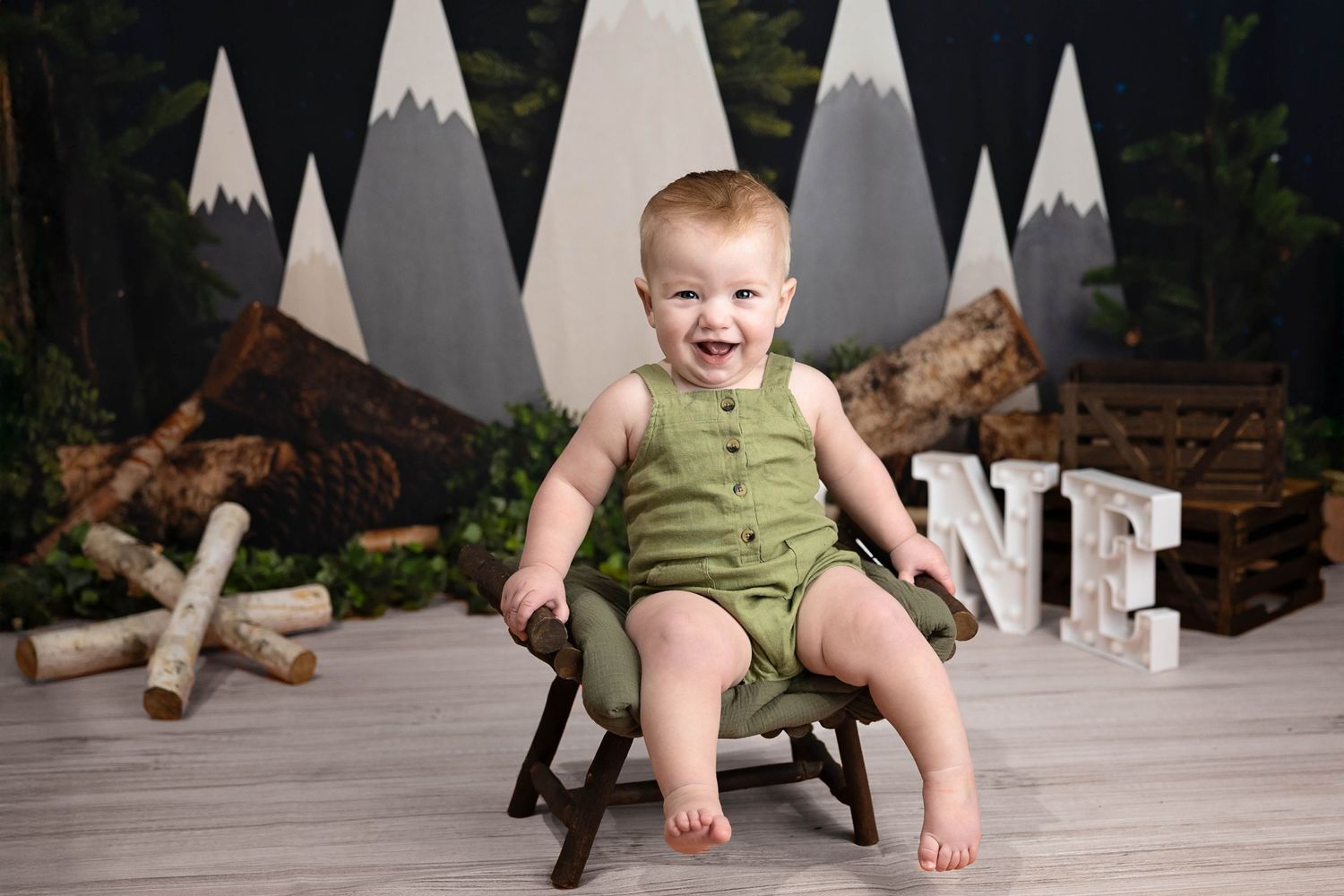 Baby in green romper sits in wooden chair against woodland-themed backdrop with painted mountains and rustic decor. Cake Smash Roswell NM