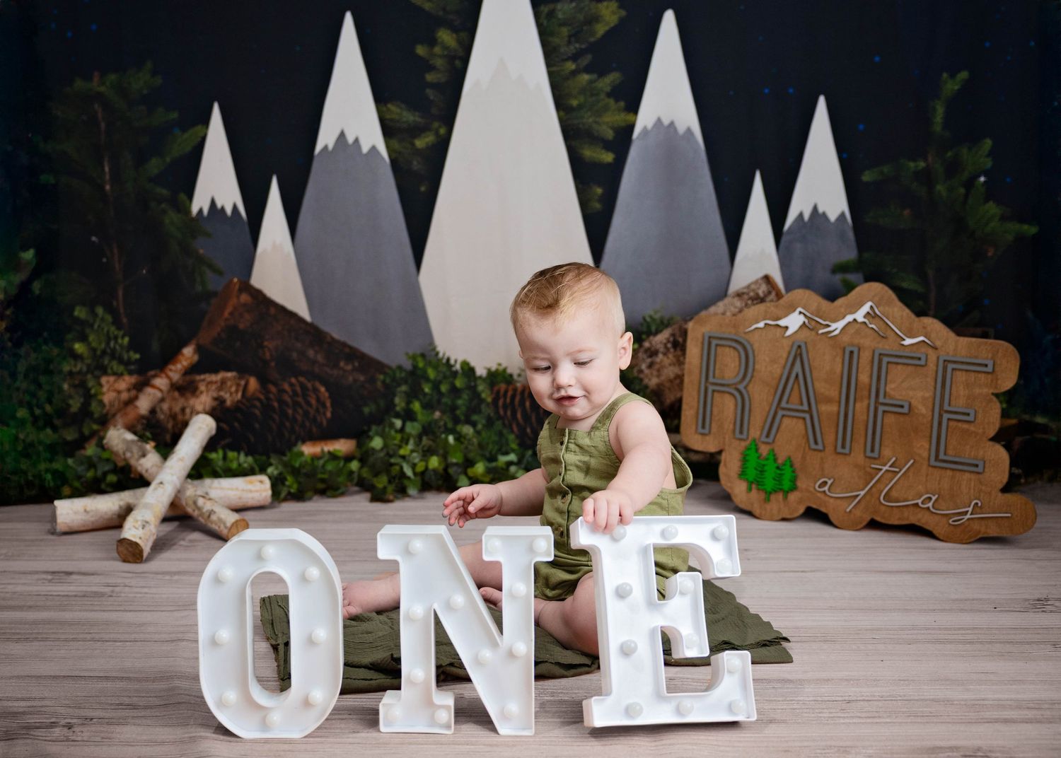 Mountain-themed first birthday photo setup with light-up 'ONE' letters and wilderness decorations against gray backdrop. Cake Smash Roswell