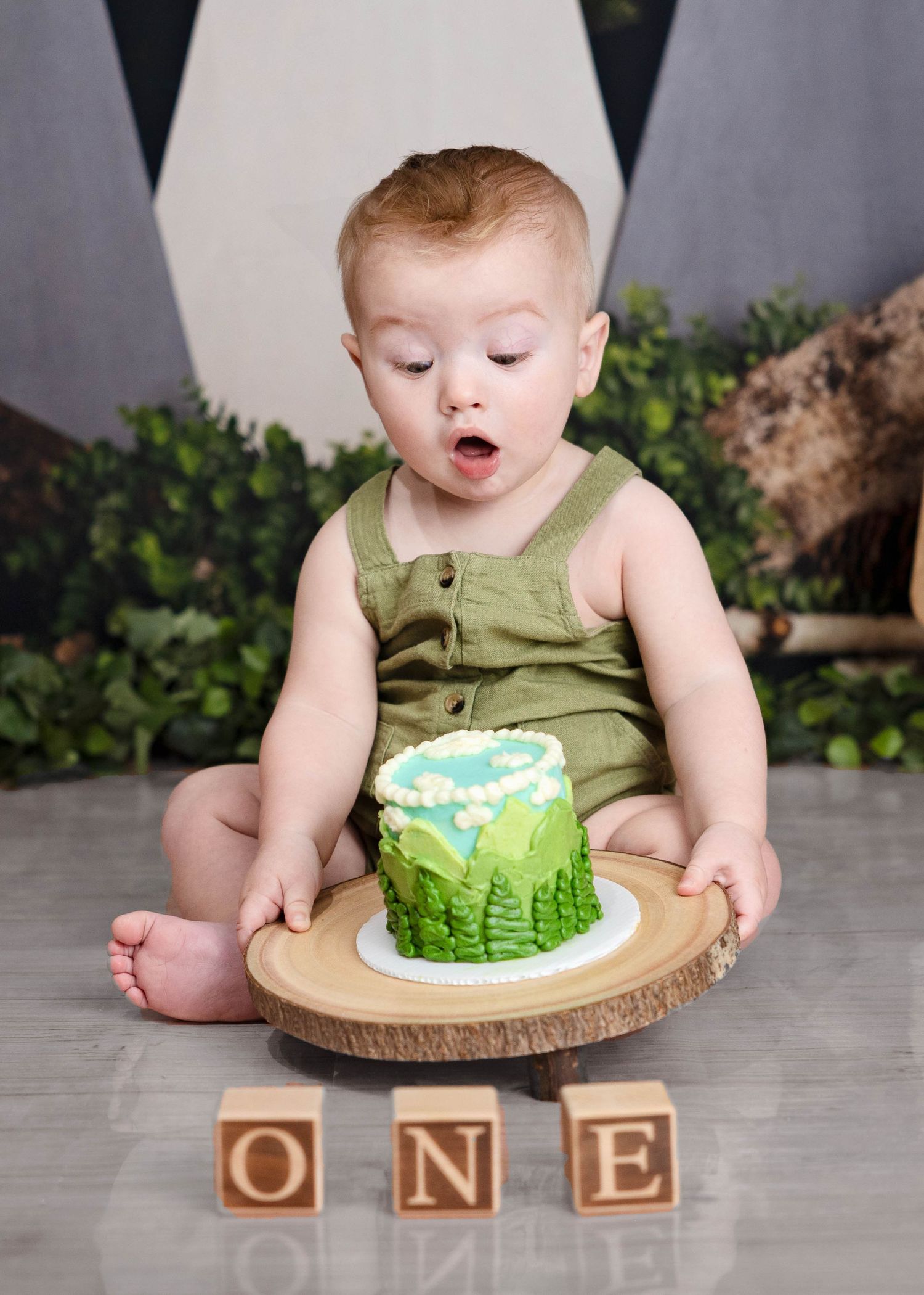 A small child in green overalls sits with their first birthday cake featuring green frosting on a wooden platter. Cake Smash Carlsbad NM
