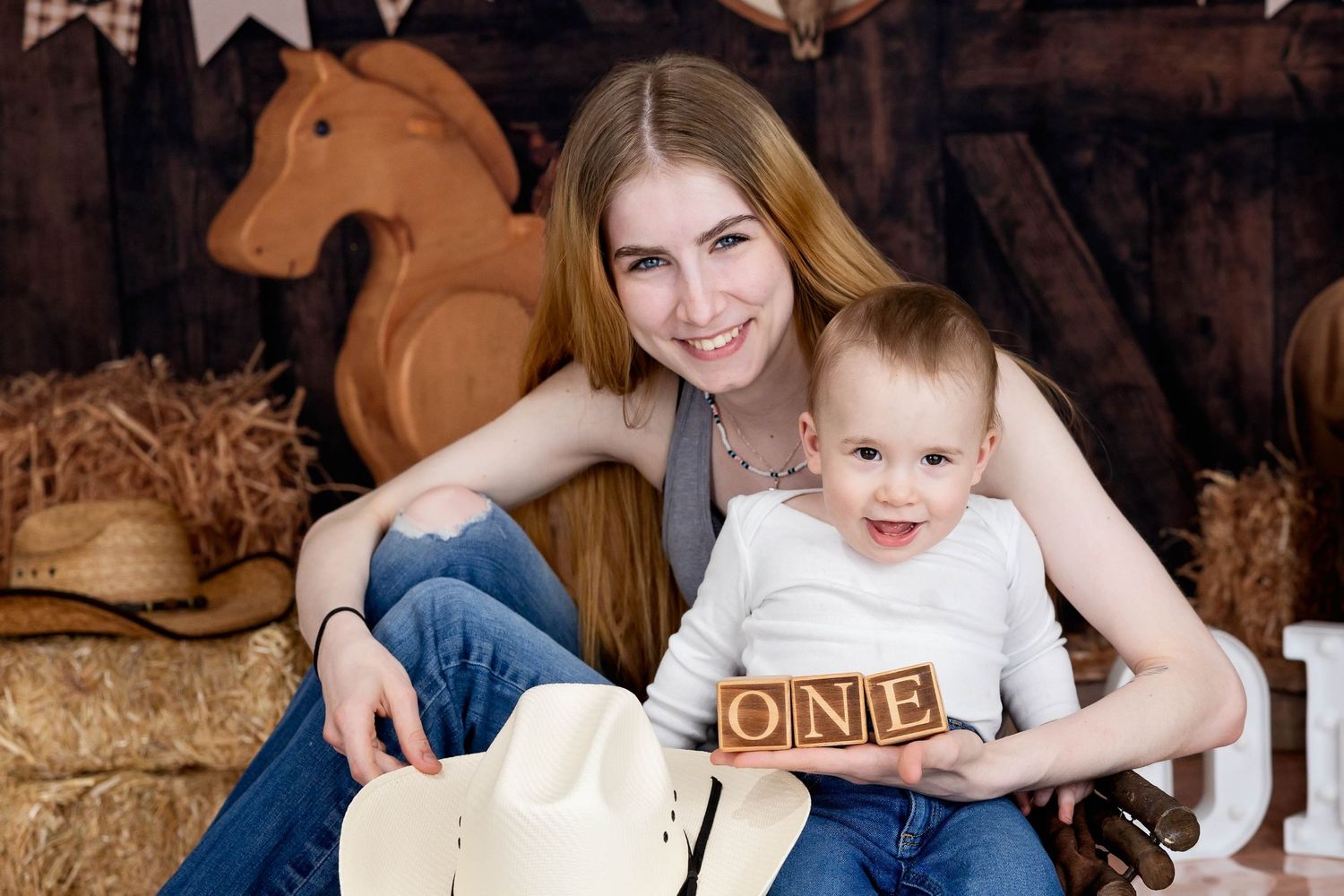 A baby holding wooden blocks reading "ONE" sits in a rustic barn setting with a wooden horse in the background. #MommyandMeBirthdayPhotos
