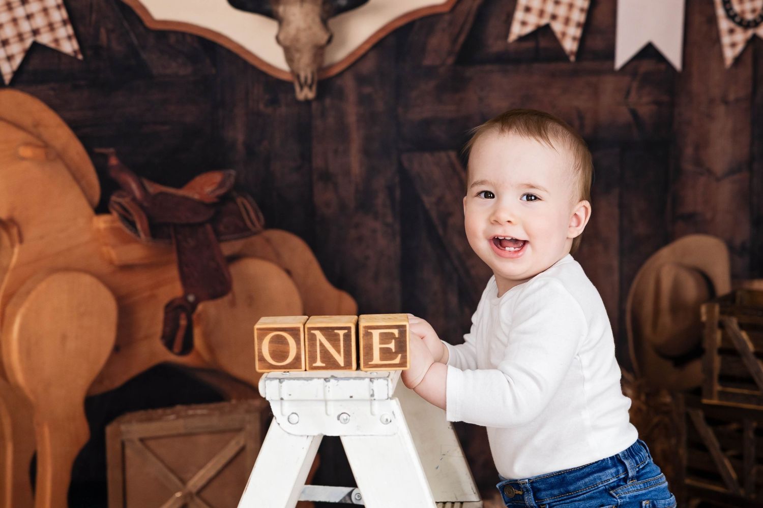 Baby in white sweater and jeans smiles while standing next to wooden blocks with "ONE" text against dark backdrop. #FirstBirthdayPhotos