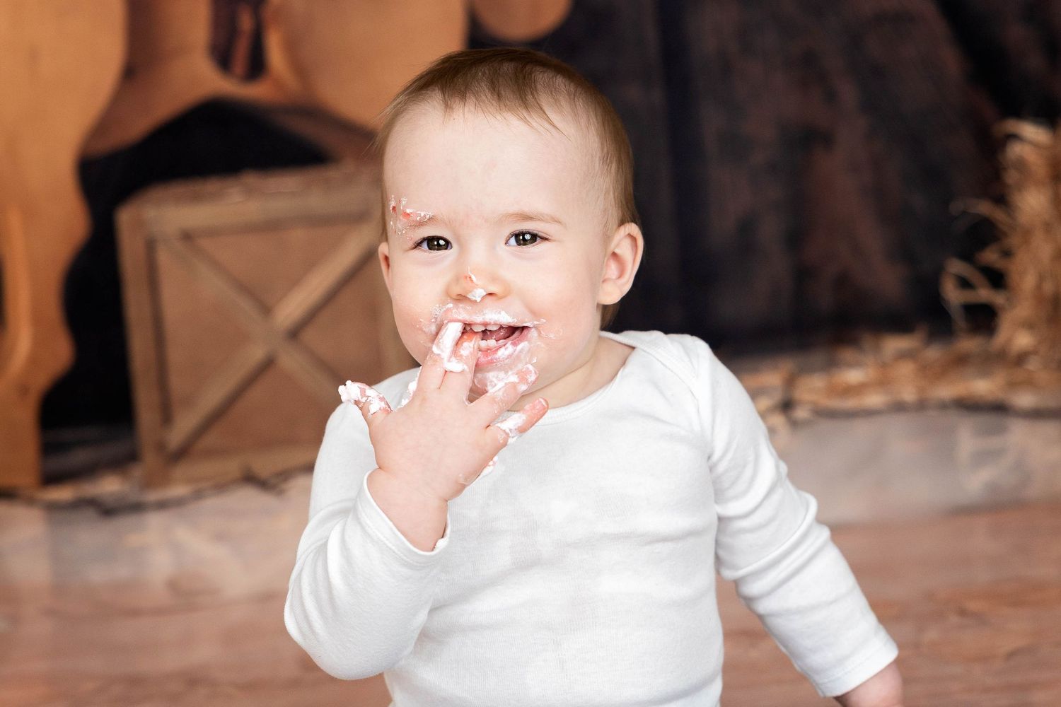 A happy baby wearing a white long-sleeve shirt smiles while sitting on a brown floor. #RoswellNM #FirstBirthday #Pictures #CarlsbadPhotos