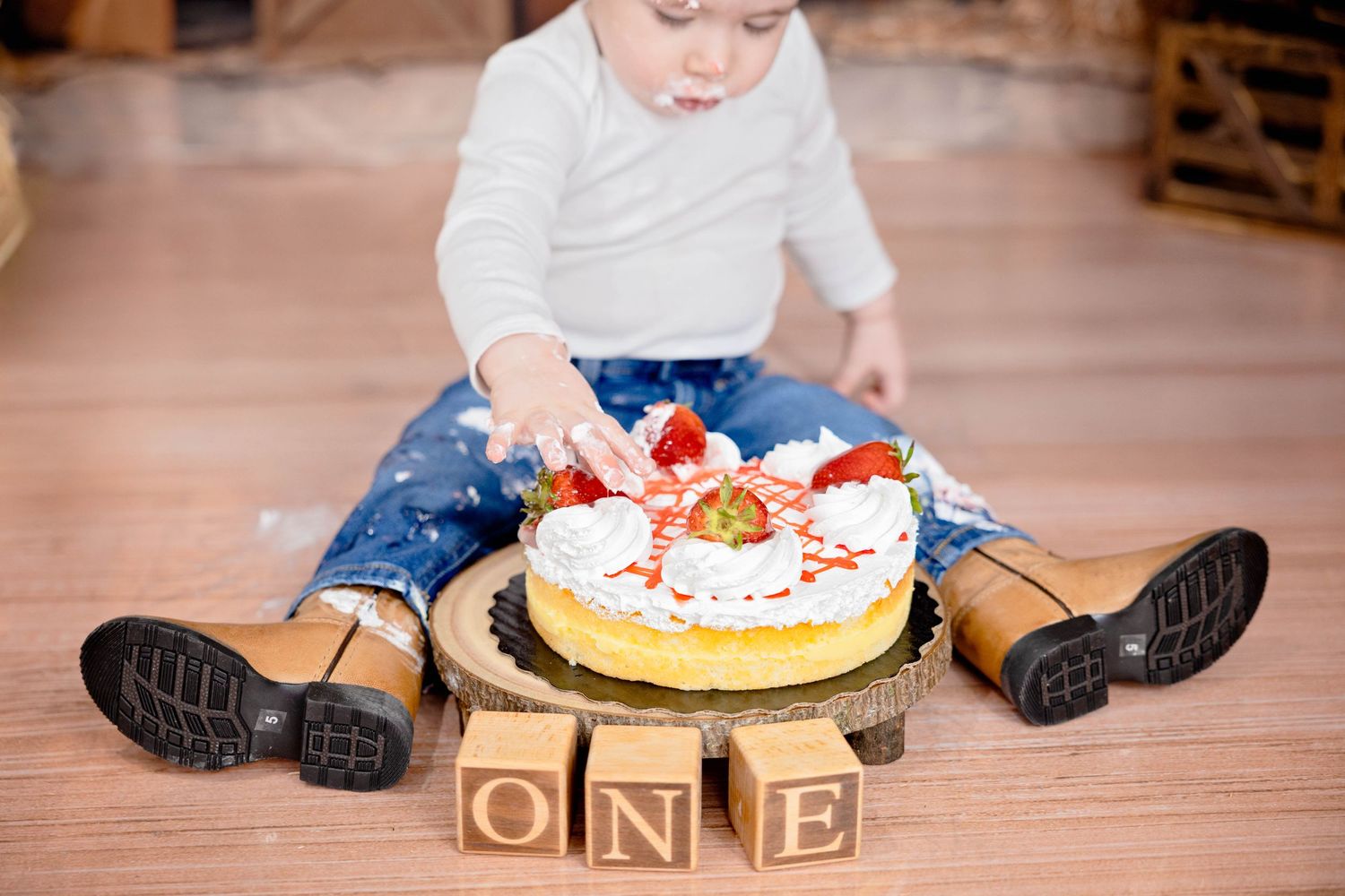 Small child in blue jeans and black boots smashes cake on wooden floor with wooden alphabet blocks spelling ONE. #CakeSmash #cowboy #Artesia