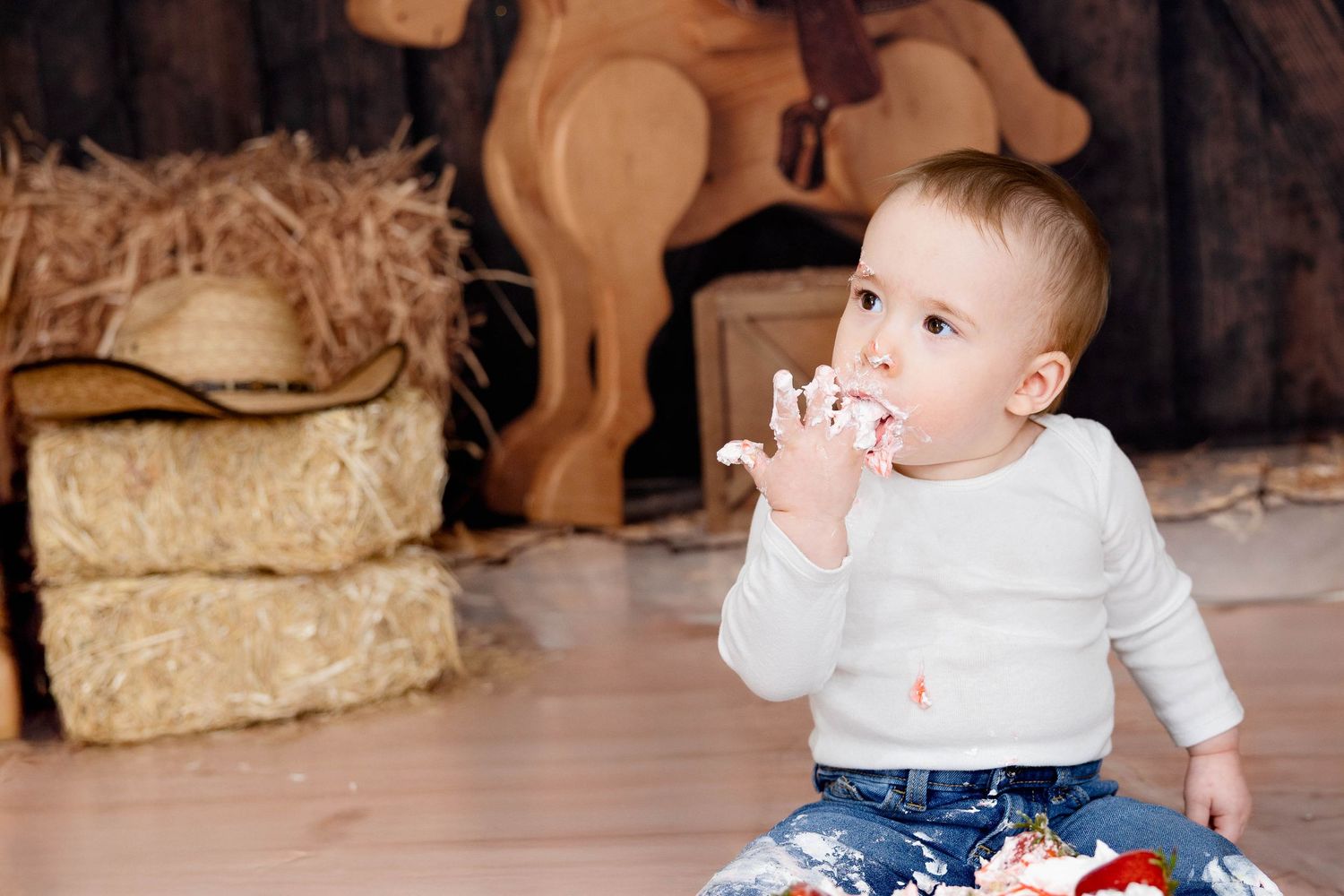 Small child sits on floor in white sweater and patterned pants with rustic decor and straw bale in background. #CakeSmash #NewMexico