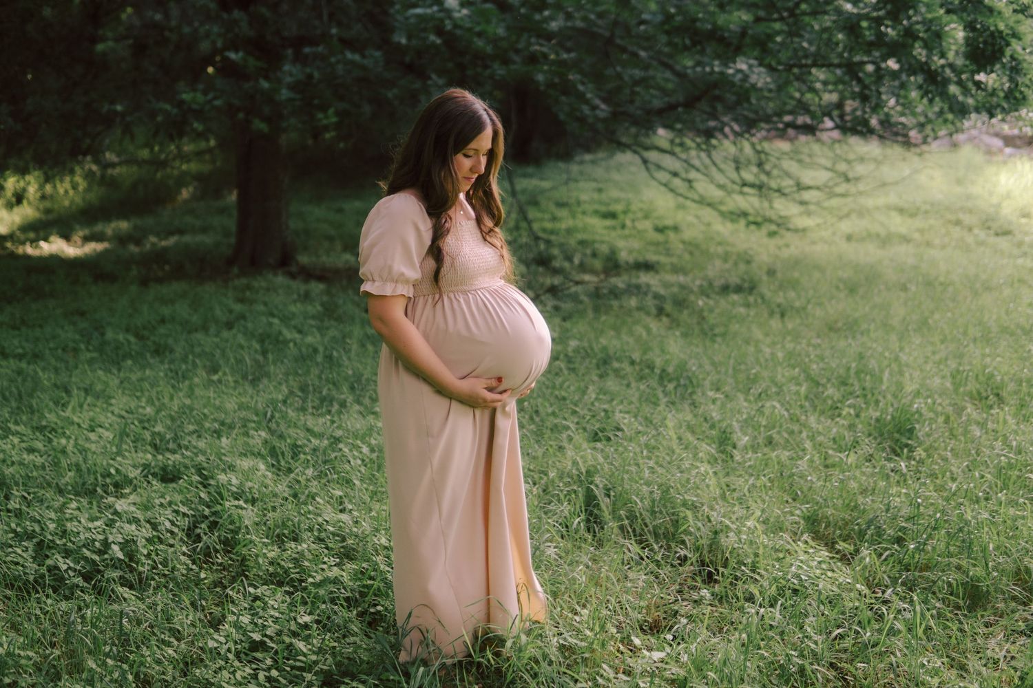An expectant mother in a blush pink maxi dress stands in a sunlit forest setting, gently holding her pregnant belly.