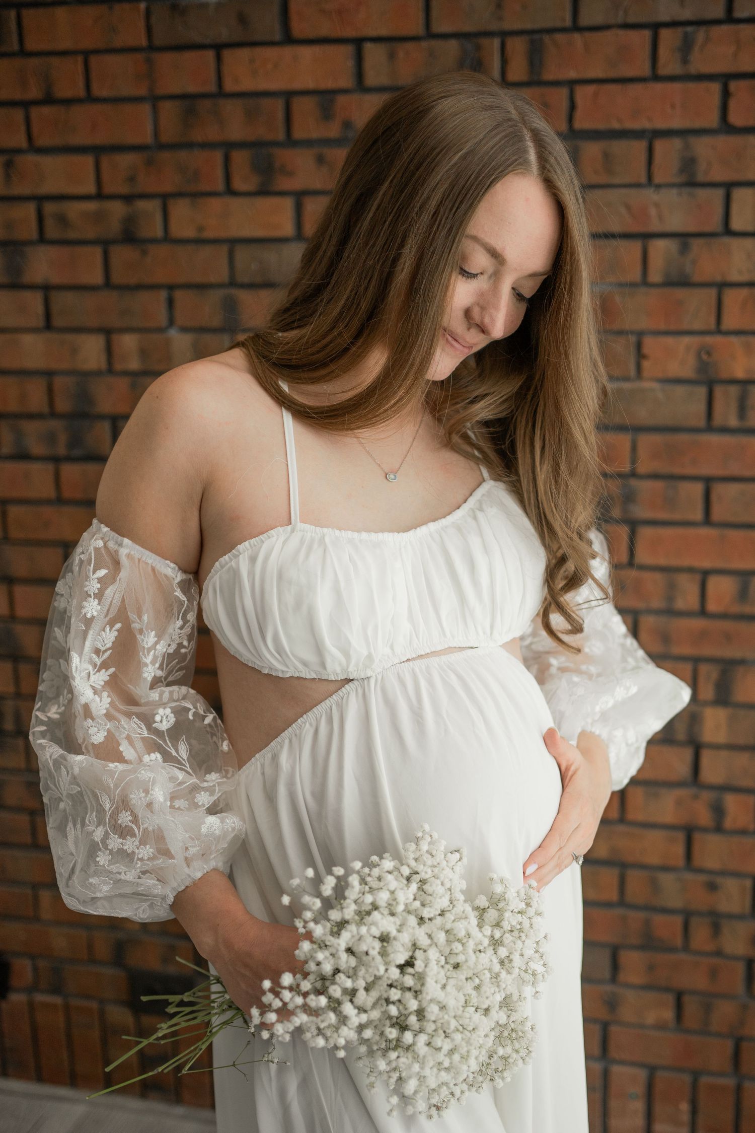 Mother-to-be in white lace dress holds baby's breath flowers against brick backdrop.