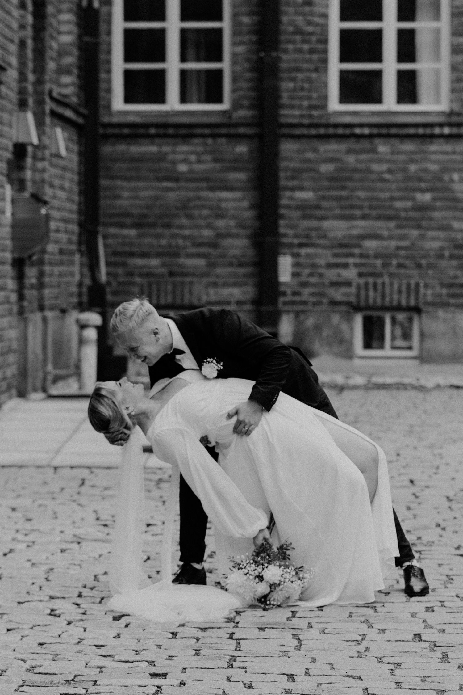 A romantic black and white wedding photo showing a classic dip pose in front of a brick building.
