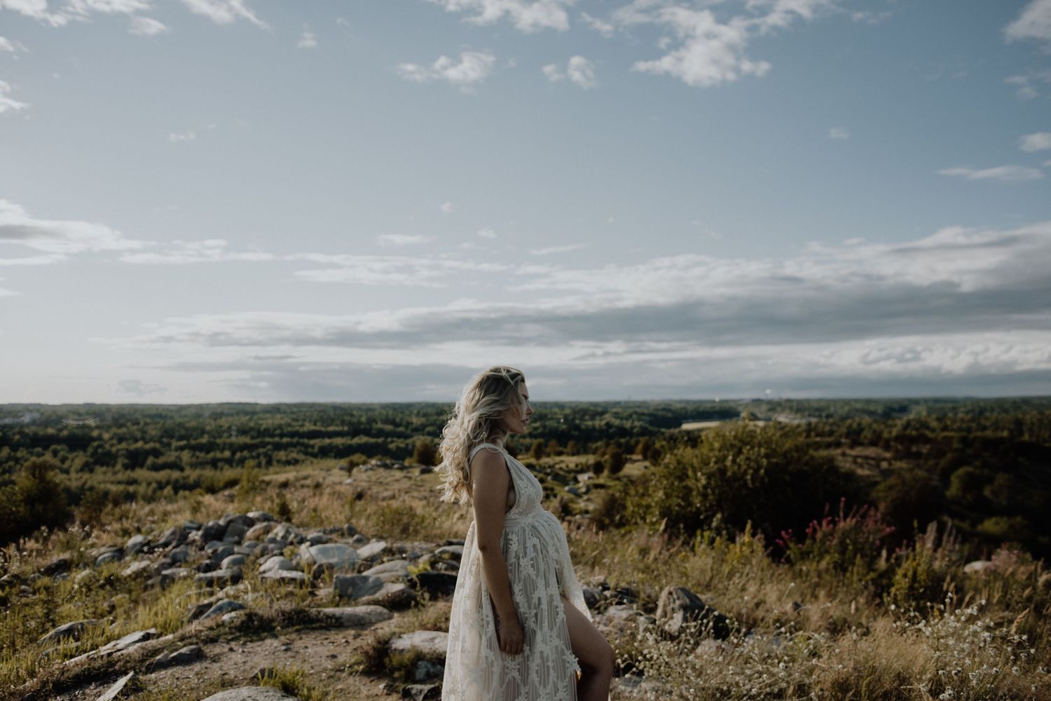 Flowing white dress catches the breeze atop a rocky hill overlooking a vast green landscape under cloudy skies.